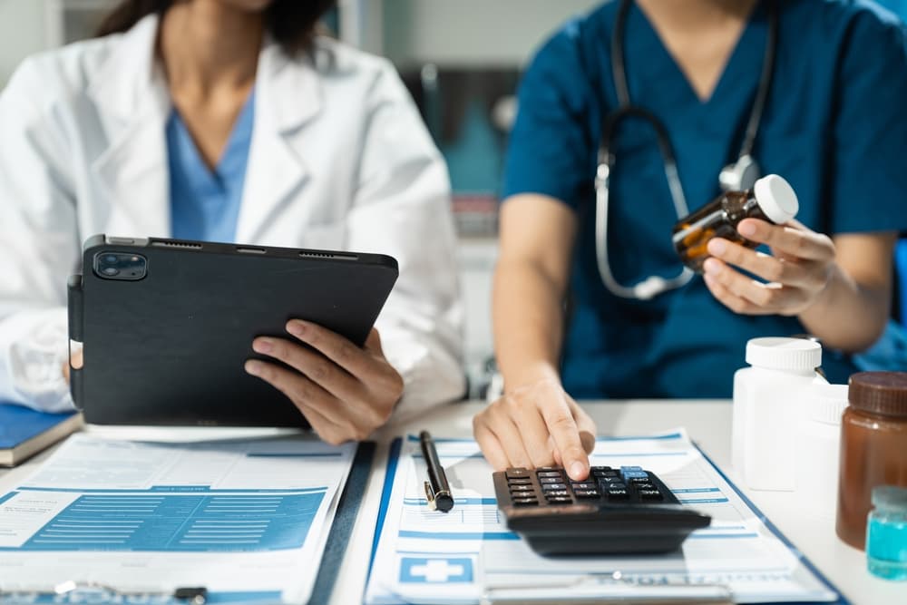 A doctor or accounting department staff writes a note while using a calculator to determine hospital staff expenses and income.