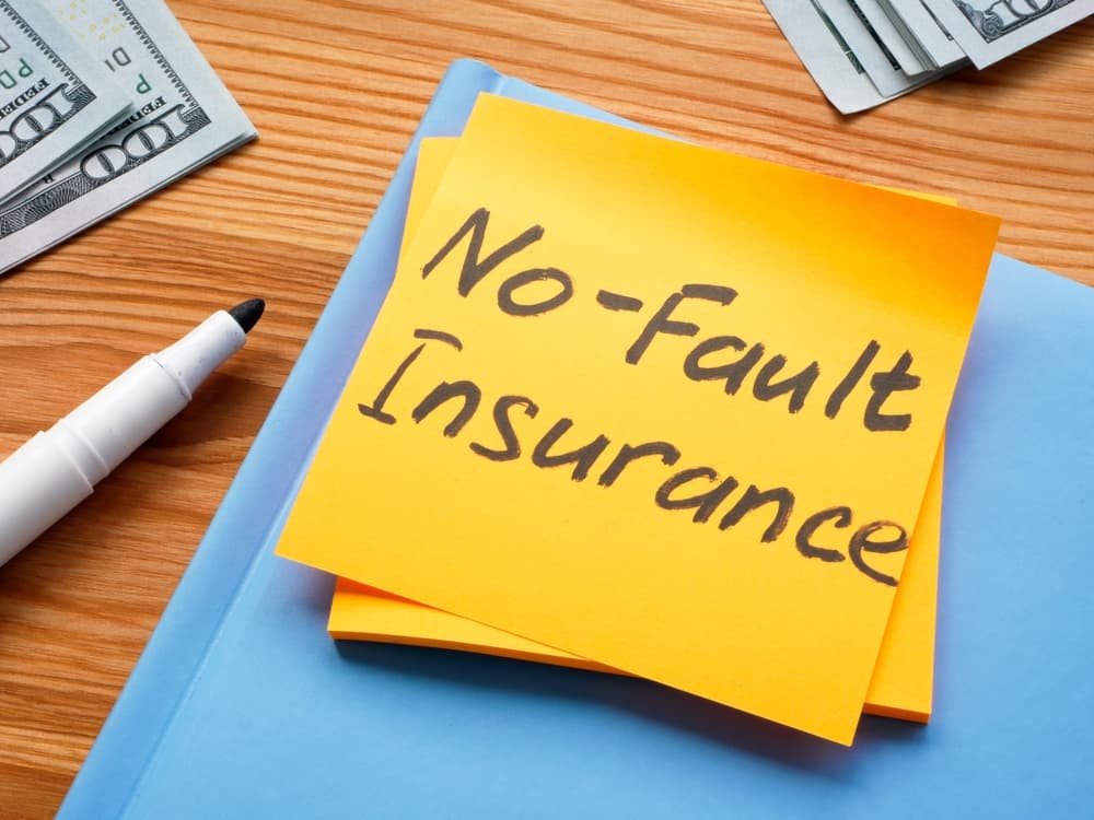 The image you requested: a notepad with a bright sticker labeled "No Fault Insurance" and a pen nearby, placed on a wooden desk in an office setting.