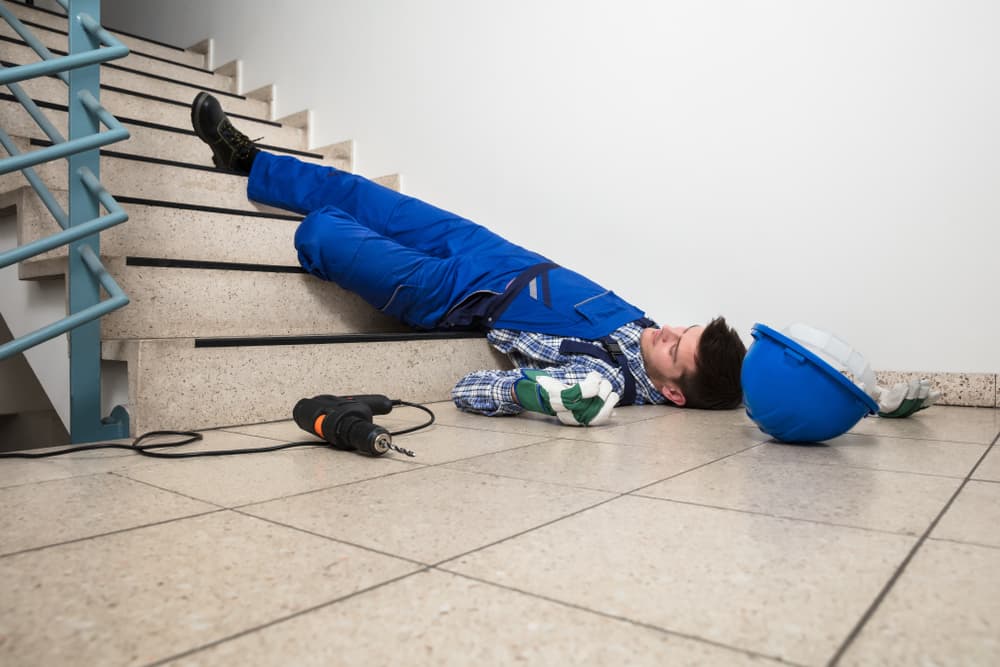 A handyman lying unconscious on a staircase, with a helmet and drill on the floor nearby.