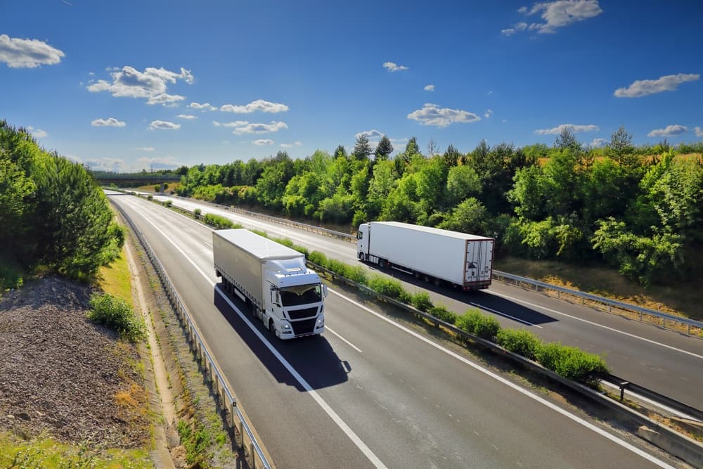 A cargo truck drives along the highway at sunset, moving through a scenic landscape with warm, golden hues casting long shadows across the road.