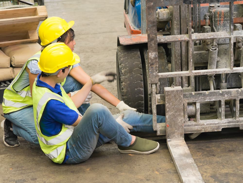 An Asian engineer was crushed by a forklift in a factory warehouse, trapping his leg. A female engineer rushes to help the injured worker.