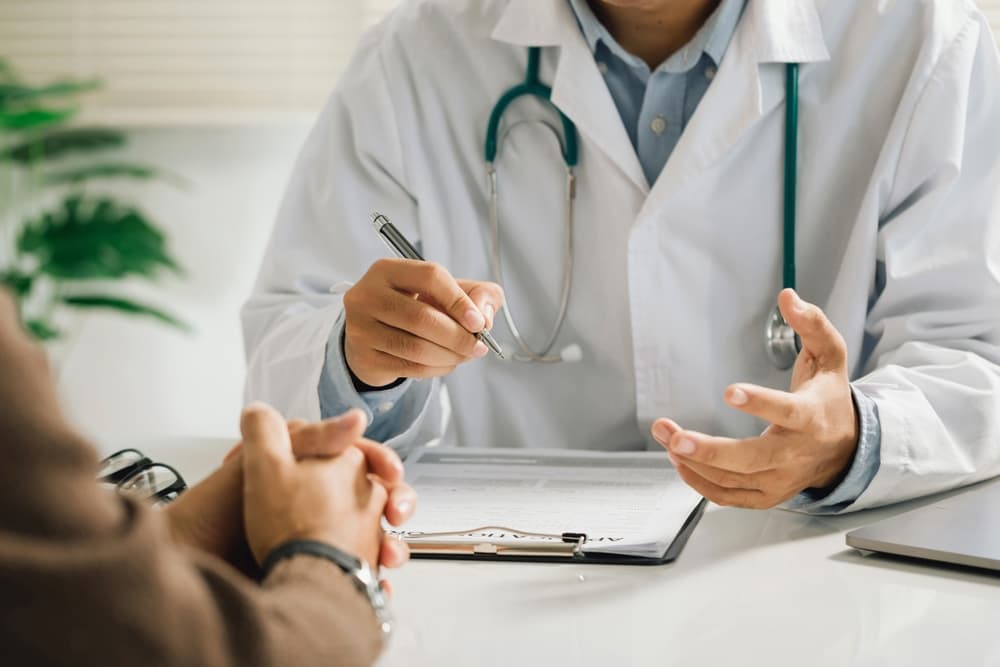 A professional physician in a white medical uniform discusses symptoms or test results with a male patient, provides recommendations, and signs a medical document during a clinic appointment.