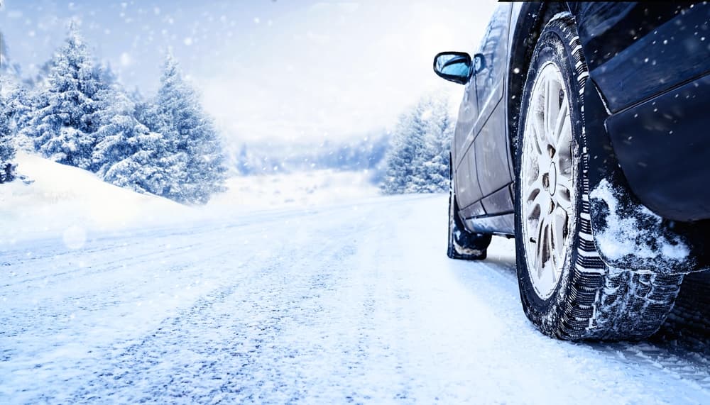 Close-up of winter tires on a car driving on a snowy road with a stunning winter landscape in the background.