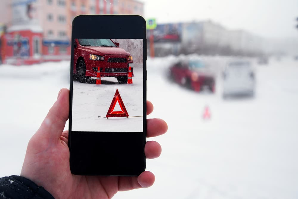 Close-up of a hand holding a smartphone, capturing a photo of a car accident on a snowy winter road.







