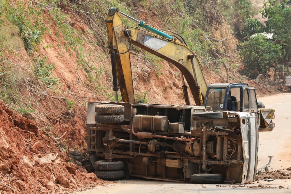 A dump truck involved in an accident at a construction site, with excavators clearing debris from the road, creating an unsafe environment.